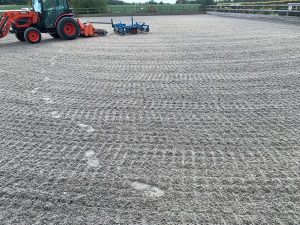Riding arena with gravel footing; a red tractor and blue equipment sit near the fence at the edge of the space.