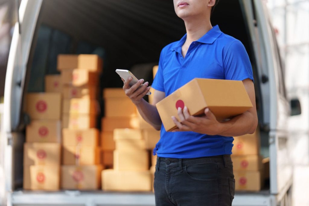 Delivery person in a blue polo holds a cardboard box and checks a smartphone in front of an open van filled with packages.